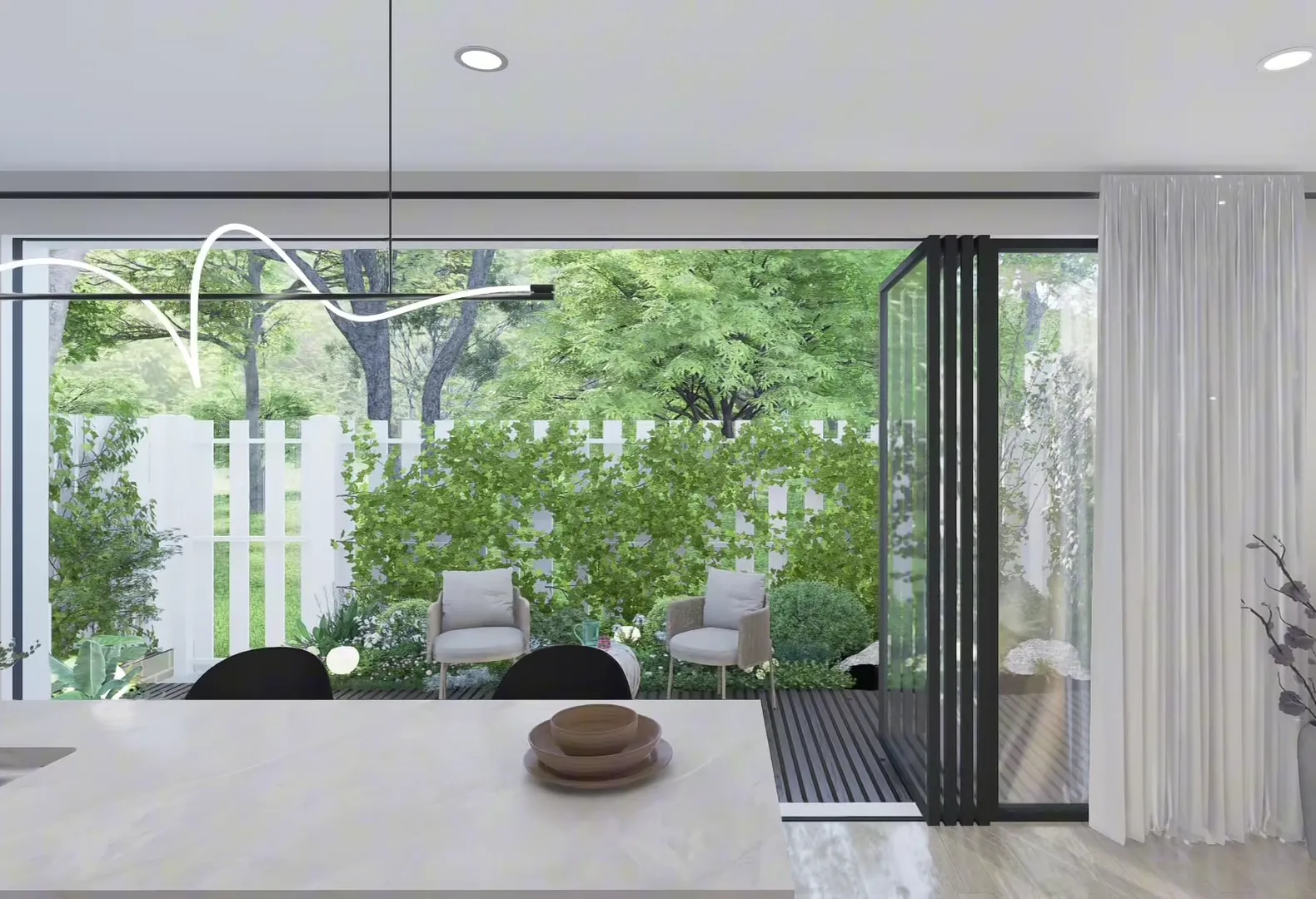 A stone kitchen island with a curved pendant above it, looking through folded-open stacker doors to a decked courtyard with lounge chairs, climbing greenery and a white picket fence.