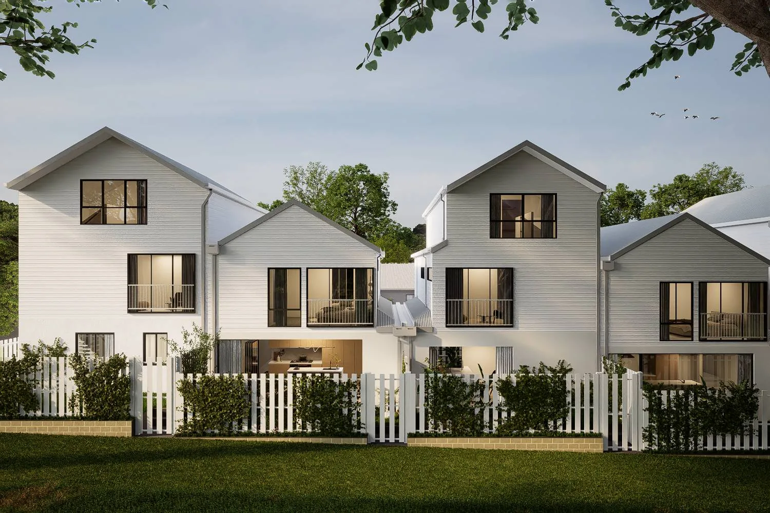 Front elevation of the townhouses at dusk, seen across the lawn of Mills Avenue Reserve, with white weatherboard gables, lit interiors behind black-framed windows and a white picket fence.