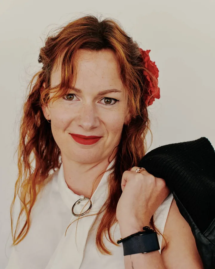 Studio portrait of a woman with long red hair and a red flower at her temple, a jacket held over one shoulder.